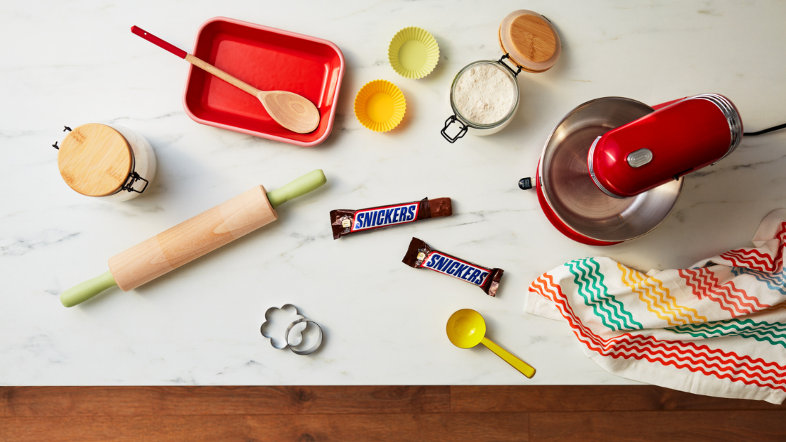 Snickers bars on a kitchen counter with baking utensils and ingredients