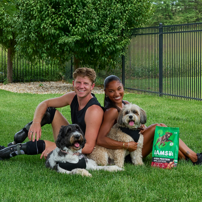Gold medalists Tara Davis-Woodhall and Hunter Woodhall sit on the grass with their dogs, Milo and Winnie.