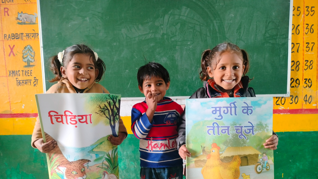Three young Indian children holding up signs in front of a classroom blackboard