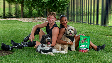 Gold medalists Tara Davis-Woodhall and Hunter Woodhall sit on the grass with their dogs, Milo and Winnie.
